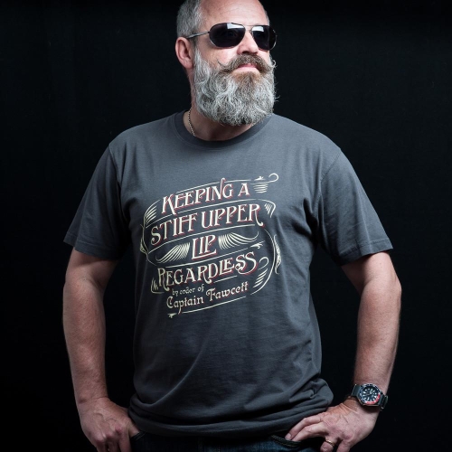 Grafitowy T-shirt z napisem Keeping a stiff upper lip regardless by order of Captain Fawcett, mężczyzna z siwą bródką i okularami, krótki rękaw, luźny krój, ciemne tło, ręka na biodrze.