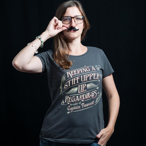 Szara koszulka z krótkim rękawem i czerwono-białym nadrukiem z hasłem Keeping a Stiff Upper Lip... Modelka w okularach, z falowanymi włosami, trzyma czarny wąsik na palcu. Tło ciemne, jednolite.