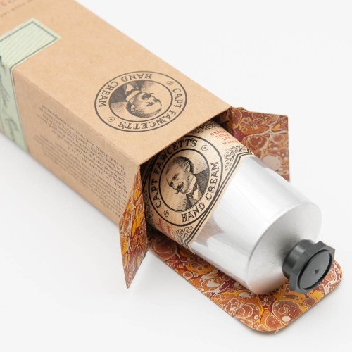 Krem do rąk w metalowej tubie z czarną nakrętką, częściowo w brązowym kartonie z pomarańczowo-złotym wzorem, z etykietą CAPT FAWCETT'S HAND CREAM i portretem postaci na białym tle.