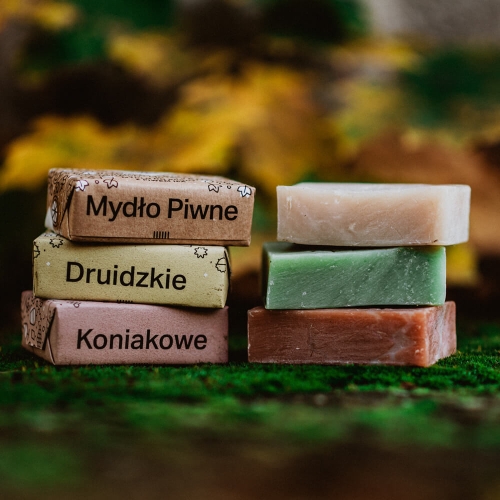 Trzy mydła: brązowe Mydło Piwne, zielone Druidzkie i różowe Koniakowe ułożone w stosie przy zielonym podłożu, na tle żółto-brązowym przypominającym naturalne środowisko.