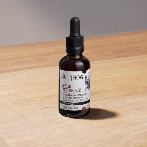 Butelka olejku do brody Bullfrog Secret Potion N°2, 50 ml, ciemnobrązowy flakon z czarną zakrętką i czerwoną etykietą z nazwą i informacjami w języku włoskim i angielskim, stoi na drewnianym blacie.