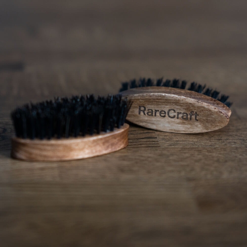 Dwie okrągłe szczotki do beard care z brązowym drewnianym uchwytem i czarnym włosiem, widoczne z boku na drewnianym tle. Jedna szczotka częściowo zasłania drugą. Na uchwycie jednej z szczotek znajduje się napis RareCraft.