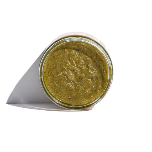 Słoik z zielonym pesto z widocznym gładkim wnętrzem, umieszczony pod kątem, na białym tle. Dostrzegalne świeże zioła i przyprawy w składzie. Słoik ma metalową zakrętkę i okrągłą formę.